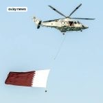 A military helicopter flying in clear blue sky carries a large Qatar flag suspended below it, with crew members visible at the open side door.