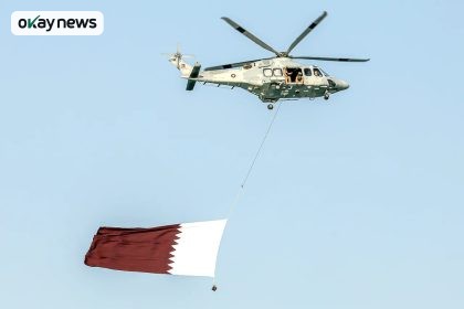 A military helicopter flying in clear blue sky carries a large Qatar flag suspended below it, with crew members visible at the open side door.