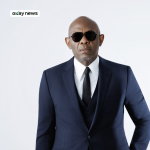 Portrait of Tony Elumelu wearing a dark blue suit, white shirt, black tie, and sunglasses, standing against a plain light background.