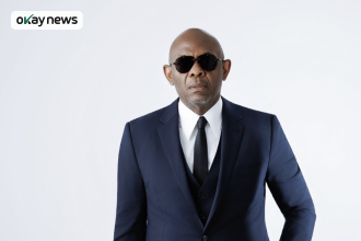 Portrait of Tony Elumelu wearing a dark blue suit, white shirt, black tie, and sunglasses, standing against a plain light background.